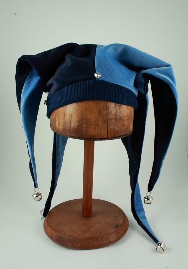 A blue and navy velvet jester hat with silver bell tips, displayed on a wooden mannequin head against a light gray background.