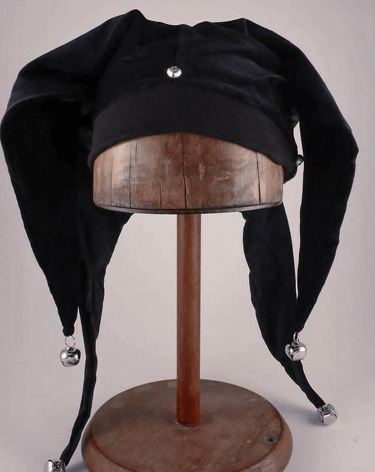 A black velvet jester hat with silver bells at the tips of its long tails, displayed on a wooden stand against a light gray background.