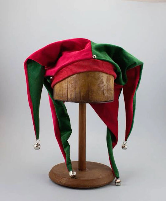 A red and green jester hat with silver bells at the ends of its long points, displayed on a wooden mannequin head stand against a light gray background.